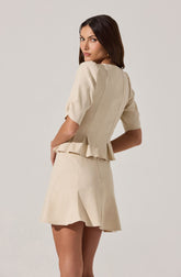 A woman is wearing a light beige outfit consisting of a short-sleeved, pleated top with puffed sleeves and a matching skirt. The top has a fitted bodice with vertical seams and a peplum hem that flares slightly at the waist. The skirt is high-waisted, slightly flared, and above knee-length.